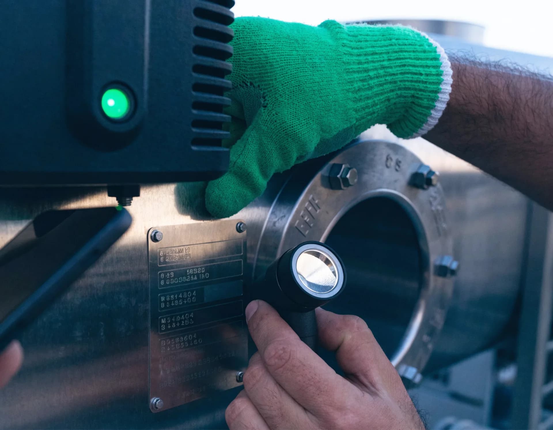 A technician's hands shine a work light on the stamped model and serial nameplate of a commercial refrigeration unit, a green status LED glowing nearby