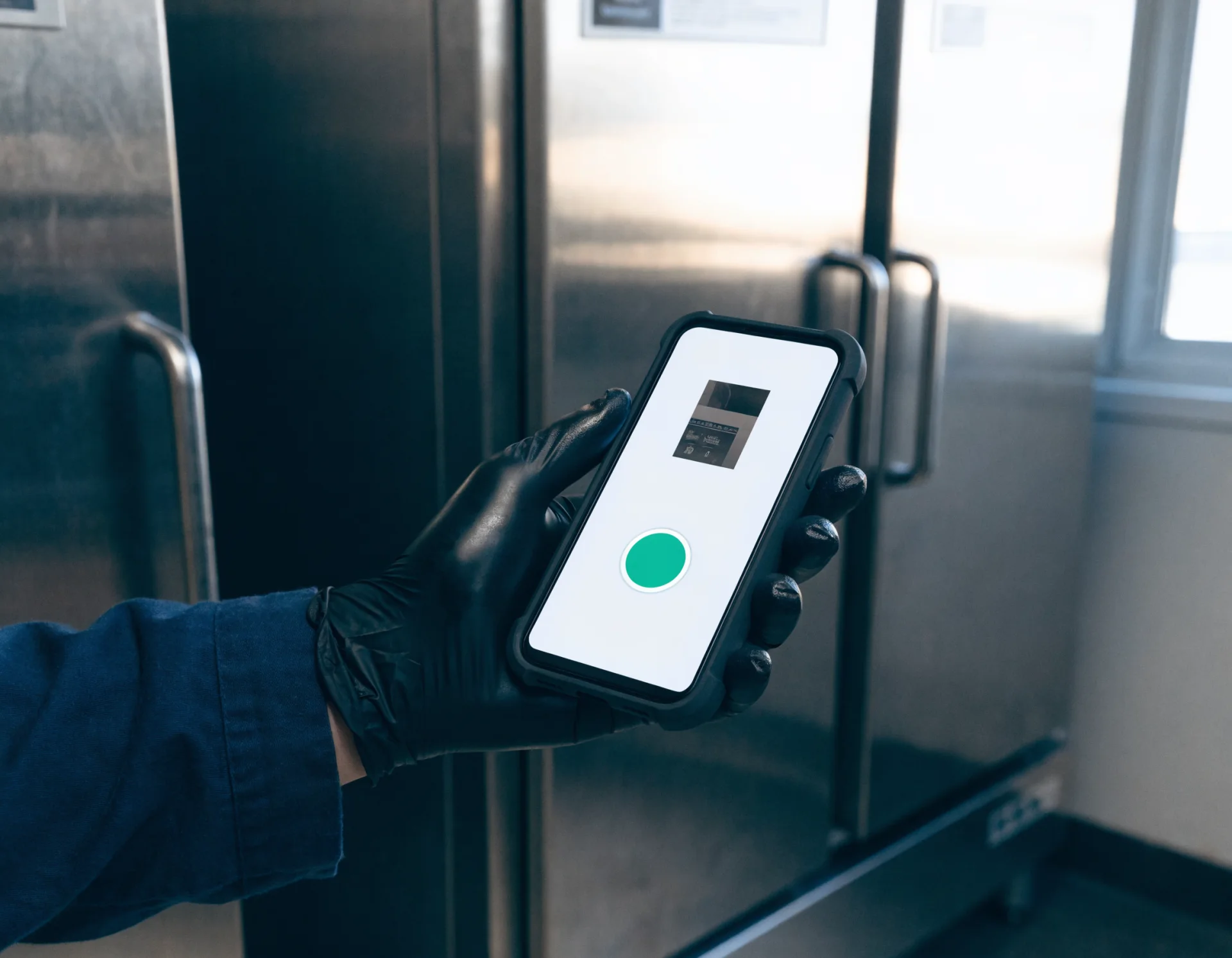 A gloved hand holds a phone showing the one-step capture app with a green camera button, in front of stainless reach-in refrigerators